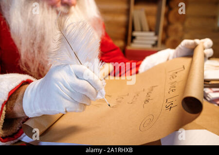 Behandschuhte hand von Santa mit White plume über schöne Liste auf Papier abgerollt, während sich bereit für Weihnachten Stockfoto