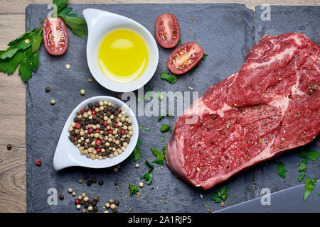 Raw Entrecote Rind Steak mit Pfeffer, Olivenöl, Knoblauch, Petersilie, Tomaten und ein Messer auf einem schwarzem Schiefer und Holz- Hintergrund Stockfoto