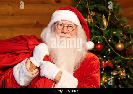 Santa Claus in traditioneller Tracht, weiße Handschuhe und Brille mit großen roten Sack voller Weihnachtsgeschenke für Kinder Stockfoto