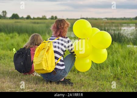 Zwei Mädchen mit Ballons Rucksäcke zurück in die Natur, Kinder in der Nähe des Sees auf der Wiese, Sommer Abend Stockfoto