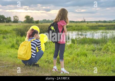 Zwei Mädchen mit Ballons Rucksäcke zurück in die Natur, Kinder in der Nähe des Sees auf der Wiese, Sommer Abend Stockfoto