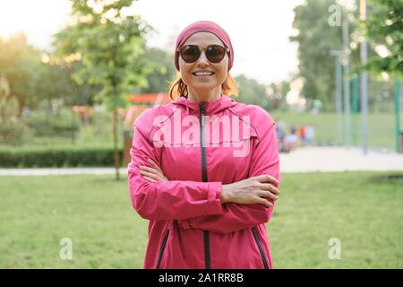 Portrait von sportlichen Reifen positive selbstbewusste Frau mit verschränkten Armen im Park in Sportkleidung Stockfoto