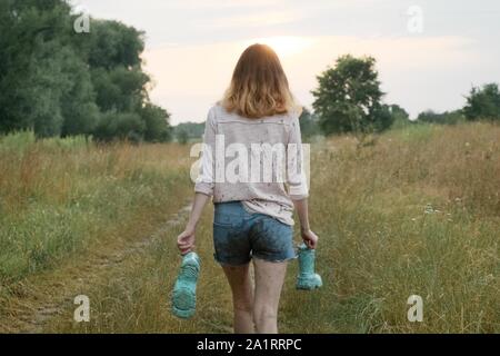 Jugendlich Mädchen gehen auf die ländliche Straße mit Stiefeln in den Händen. Happy girl in schmutziger Kleidung, Rückansicht, Sommer Natur Hintergrund Stockfoto