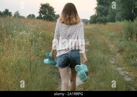 Jugendlich Mädchen gehen auf die ländliche Straße mit Stiefeln in den Händen. Happy girl in schmutziger Kleidung, Rückansicht, Sommer Natur Hintergrund Stockfoto