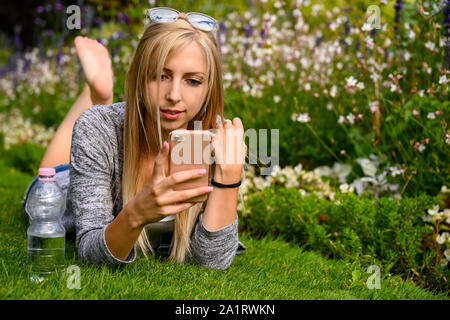 Passende Frau mit Trinkwasser in einer Flasche mit ihrem Mobiltelefon Kontrolle in einem Park oder Garten in London, Vereinigtes Königreich Stockfoto