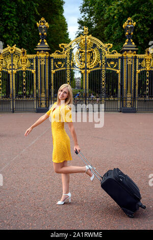 Attraktive junge Frau in gelb Fancy Dress, ziehen einen kleinen Koffer und lächelnd und am Buckingham Palace, London, Vereinigtes Königreich, Stockfoto