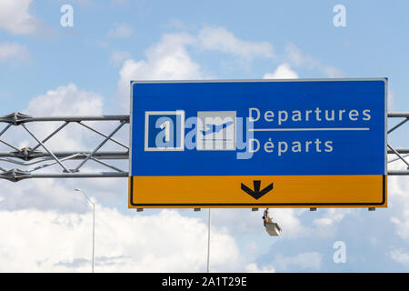 Schild für die Flughafenabflüge (Englisch/Französisch) über der Fahrbahn an teilweise bewölktem Tag. Stockfoto