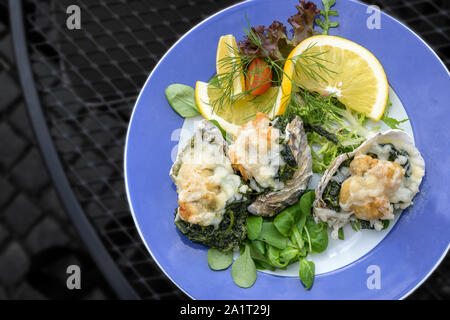 Gebackene Austern Rockefeller überbacken mit Parmesan und Spinat, mit Salat auf einem blauen Platte auf dunklem Metall Tisch serviert, hohe Betrachtungswinkel von ab Stockfoto