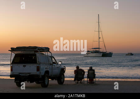 Ein paar sitzen in Liegestühlen neben einem Allradantrieb wie die Sonne über dem Indischen Ozean am Cable Beach in Broome, Western Australia. Stockfoto