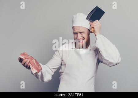 Portrait von attraktiven stattlichen Metzger guy Holding frisches Fleisch und Messer in die Arme mit den Händen berühren Kopf clueless Heiß kochen Küche vorzubereiten, isoliert Stockfoto