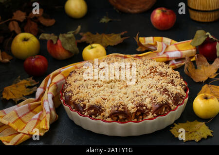 Torte mit Äpfeln wird in eine keramische Form auf einem dunklen Hintergrund entfernt, Äpfel liegen auf dem Tisch verstreut, horizontale Ausrichtung Stockfoto