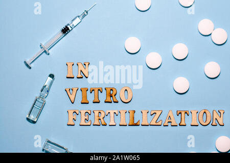 Die Inschrift In-vitro-Fertilisation ist in Holz- Buchstaben auf blauem Hintergrund. In-vitro-pen, Tablet und Ampulle. Eine Spritze mit einer dünnen Hu Stockfoto