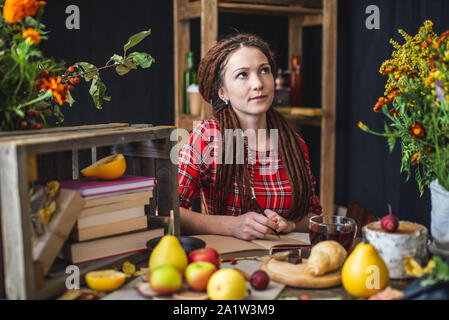 Eine schöne junge Frau in einem roten Kleid in einem Herbst Tabelle mit gelben Blumen und Kürbisse sitzen ist schriftlich in einem Notebook. Das Konzept der Herbst Rom Stockfoto