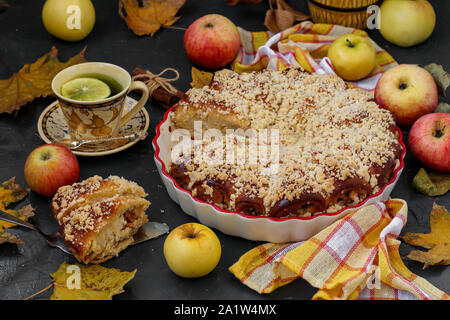 Torte mit Äpfeln wird in eine keramische Form auf einem dunklen Hintergrund entfernt, Äpfel liegen auf dem Tisch verstreut, horizontale Ausrichtung Stockfoto