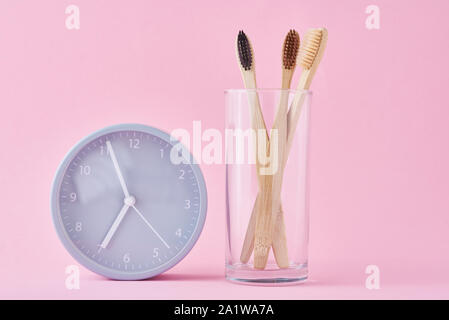 Drei Holz Bambus Zahnbürsten in Glas und Wecker auf rosa Hintergrund. Morgen Hygiene, Zahnpflege Konzept Stockfoto