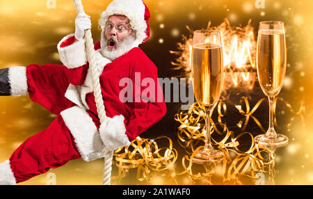 Frohe Weihnachten Santa Claus Klettern am Seil mit Gläser Champagner in der Hintergrund. Kaukasische männliches Modell in der traditionellen Tracht. Konzept der Feiertage, Silvester, winter Stimmung, Geschenke. Copyspace. Stockfoto