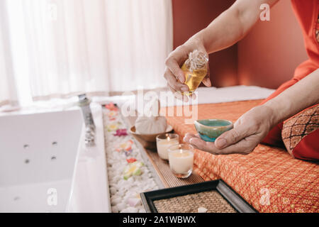 7/8-Ansicht von Masseur sitzen auf Massage Matte und Gießen Duft Öl zur Schüssel Stockfoto