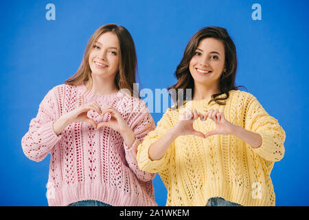 Attraktive und lächelnde Frauen in Pullover zeigen Herz Geste isoliert auf Blau Stockfoto