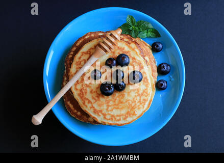 Stapel von hausgemachte Pfannkuchen auf türkis blaue Platte mit Honig, Blaubeeren und mit Puderzucker bestäubt. Der dunkle Hintergrund. Ansicht von oben Stockfoto