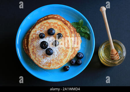 Stapel von hausgemachte Pfannkuchen auf türkis blaue Platte mit Honig, Blaubeeren und mit Puderzucker bestäubt. Der dunkle Hintergrund. Ansicht von oben Stockfoto