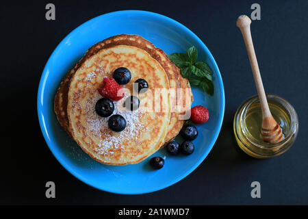 Stapel von hausgemachte Pfannkuchen auf türkis blaue Platte mit Honig, Blaubeeren und mit Puderzucker bestäubt. Der dunkle Hintergrund. Ansicht von oben Stockfoto