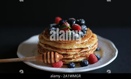 Stapel Pfannkuchen mit Honig, Blaubeeren und roten Himbeeren bestäubt mit Puderzucker auf einem dunklen Hintergrund Stockfoto