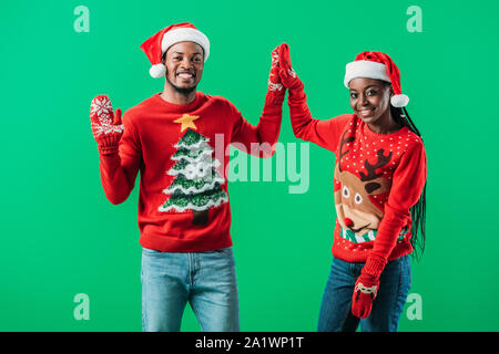 Afrikanische amerikanische Paar in Red Christmas Santa Hüte Pullover und hohe fünf untereinander und mit Blick auf die Kamera isoliert auf Grün, Stockfoto