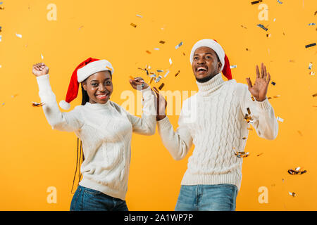 Aufgeregt Afrikanische amerikanische Ehepaar in Santa Hüte auf gelbem Hintergrund mit Konfetti Stockfoto