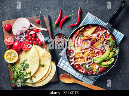 Chilaquiles mit Schinken, zerbröckelt panela Käse, Avocado und Tomaten Salsa in einer Pfanne auf einer konkreten Tabelle mit Zutaten auf einem Schneidebrett, Me Stockfoto