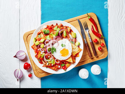 Hausgemachte chilaquiles mit einem Spiegelei, Schinken, zerbröckelt panela Käse, Avocado und Tomaten Salsa auf einer weißen Platte auf einem Schneidebrett, mexikanische Gericht, Anzeigen Stockfoto