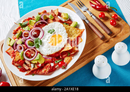 Hausgemachte chilaquiles mit einem Spiegelei, Schinken, zerbröckelt panela Käse, Avocado und Tomaten Salsa auf einer weißen Platte auf einem Schneidebrett, mexikanische Gerichte serviert. Stockfoto