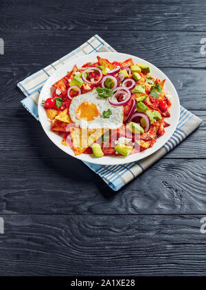 Chilaquiles mit einem Spiegelei, Schinken, zerbröckelt panela Käse, Avocado und Tomaten Salsa auf einer weißen Platte auf einem Schneidebrett, mexikanische Gericht, vertikale Ansicht Stockfoto