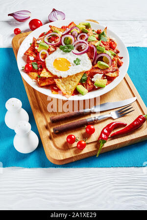 Hausgemachte chilaquiles mit einem Spiegelei, Schinken, zerbröckelt panela Käse, Avocado und Tomaten Salsa auf einer weißen Platte auf einem Schneidebrett, mexikanische Gericht, Verti Stockfoto