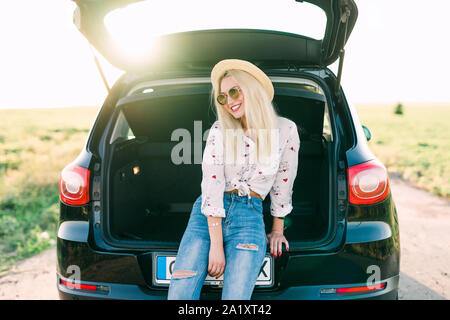 Frau sitzt im Auto lächelnd. Immer bereit zu gehen. Junge lachende Frau, die in der geöffneten Kofferraum sitzen. Sommer road trip. Junge Frau sitt Stockfoto