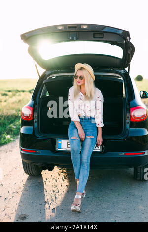 Frau sitzt im Auto lächelnd. Immer bereit zu gehen. Junge lachende Frau, die in der geöffneten Kofferraum sitzen. Sommer road trip. Junge Frau sitt Stockfoto