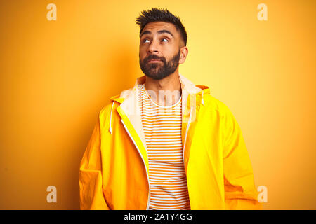 Junge indische Mann mit regenmantel stehend über isolierte gelben Hintergrund lächelnd auf die Seite und starrte weg denken. Stockfoto