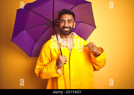 Junge indische Mann mit Regenmantel und lila Schirm über isolierte gelber Hintergrund mit Überraschung Gesicht zeigenden Finger zu sich selbst Stockfoto