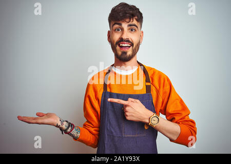 Junge Ladenbesitzer Mann mit Tattoo tragen Schürze stehend über isoliert weißer Hintergrund erstaunt und lächelt in die Kamera während der Präsentation und mit der Hand Stockfoto