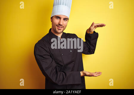 Junge Küchenchef Mann in Uniform und Mütze stehend über isolierte gelben Hintergrund und gestikulierte mit Händen zeigen große und große Zeichen, Symbol. Stockfoto