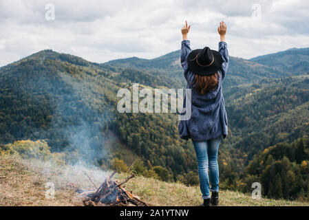 Ansicht der Rückseite des jungen Frau in hat stand in der Nähe von Lagerfeuer auf der Oberseite des Berges Gipfel, wirft die Arme in die Luft, glücklich und betrunken auf Leben, Jugend und Glück. Stockfoto