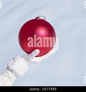 Woman's Hand mit weißen Strickhandschuh Holding groß, Red Christmas ornament Christbaumkugel mit weißen Schnee Hintergrund. Frauen kaltem Wetter Mode Accessoires. Stockfoto