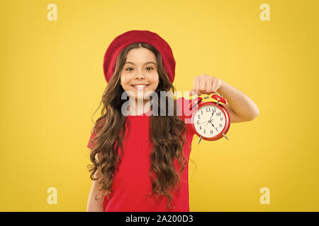 Shopping, Verkauf und Zeit Konzept. Pariser Kind auf Gelb. Kind mit Wecker. Zeitlose Mode. glückliches Mädchen mit langen Locken in Baskenmütze. Schönheit Friseur. Kleines Mädchen im französischen Stil hat. Stockfoto