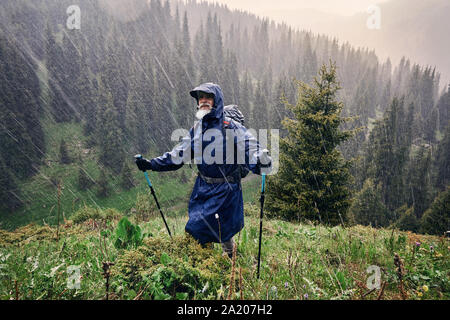 Alte Wanderer mit grauen Bart in blau Regenjacke steht unter starker Dusche in den Wald auf die Berge. Outdoor Travel Concept Stockfoto