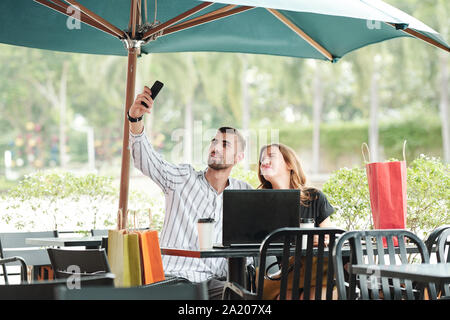 Junges Paar in Café im Freien mit geöffneten Laptop und viele Tüten herum und unter selfie sitzen zusammen Stockfoto
