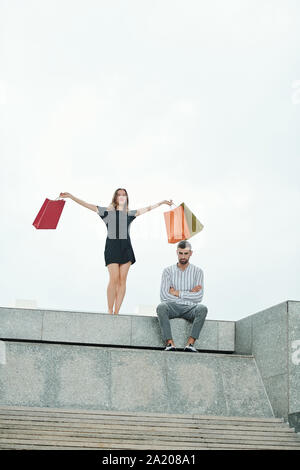 Unglückliche Menschen sitzen im Freien mit verschränkten Armen, wenn seine neugierige junge Freundin mit vielen Shopping - Taschen hinter stehend Stockfoto