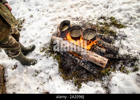 Kochen in einer Winterwanderung im Kessel über dem Feuer in der Verschneiten Kiefernwald beim Camping an einem sonnigen Tag, man steht in der Nähe von Stockfoto