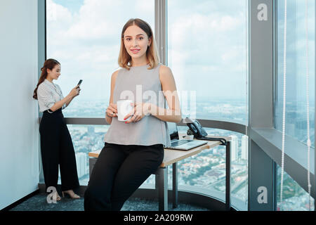 Portrait von ziemlich lächelnd weibliche Unternehmer sitzen am Rande von ihrem Schreibtisch und Kaffee trinken Stockfoto