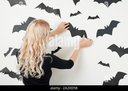 Junge Frau klemmt Fledermaus Aufkleber auf weiße Wand bei der Vorbereitung für Halloween Feier, Ansicht von der Rückseite Stockfoto