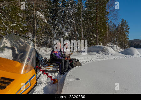 Älteres Paar einen Kaffee Bremse auf einem snowmobile Schlitten im Schnee einen sonnigen Tag im Frühjahr mit blauem Himmel und Wald im Hintergrund, Bild aus Stockfoto