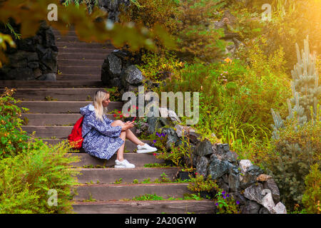 Junge schöne blonde Mädchen in einem blauen Regenmantel sitzt auf einer Holztreppe mit Steinmauern in einem gelben Herbst blühenden Garten Textnachrichten auf soziale Netzwer Stockfoto